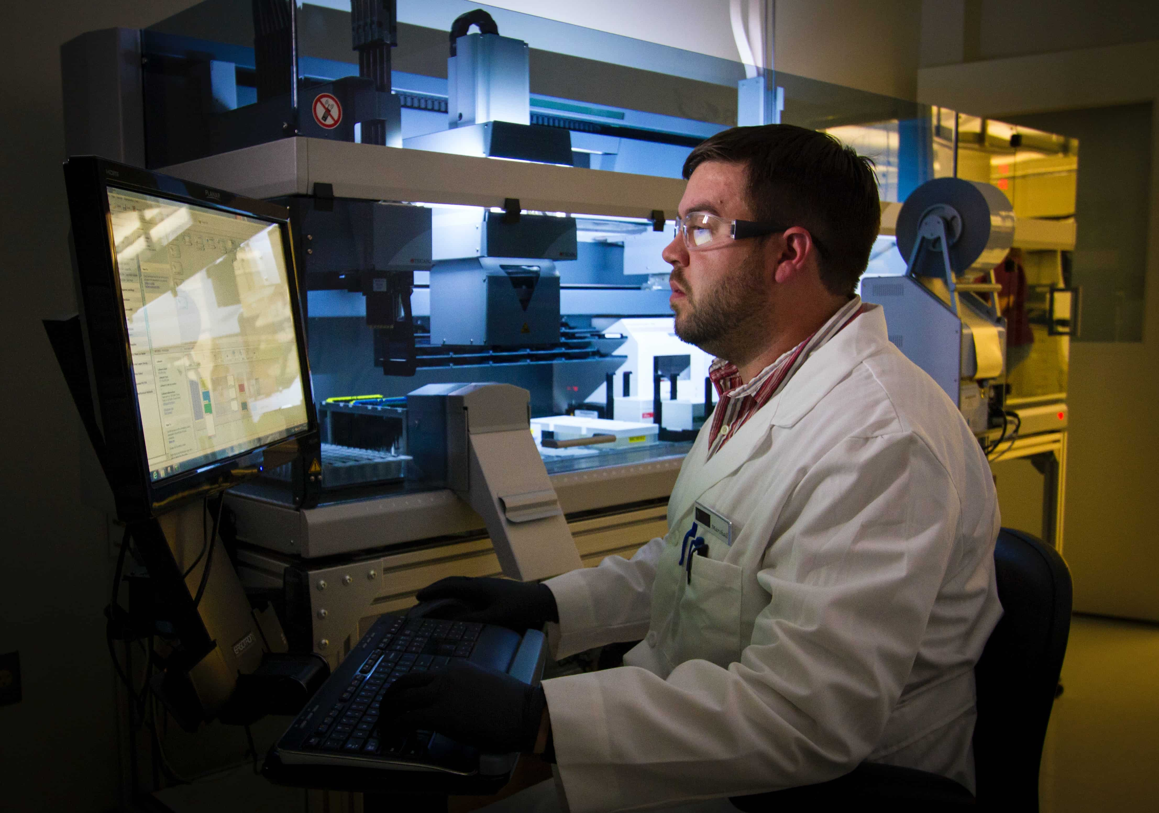 person wearing lab coat and safety glasses sitting down at screen in the background there is a scientific lab